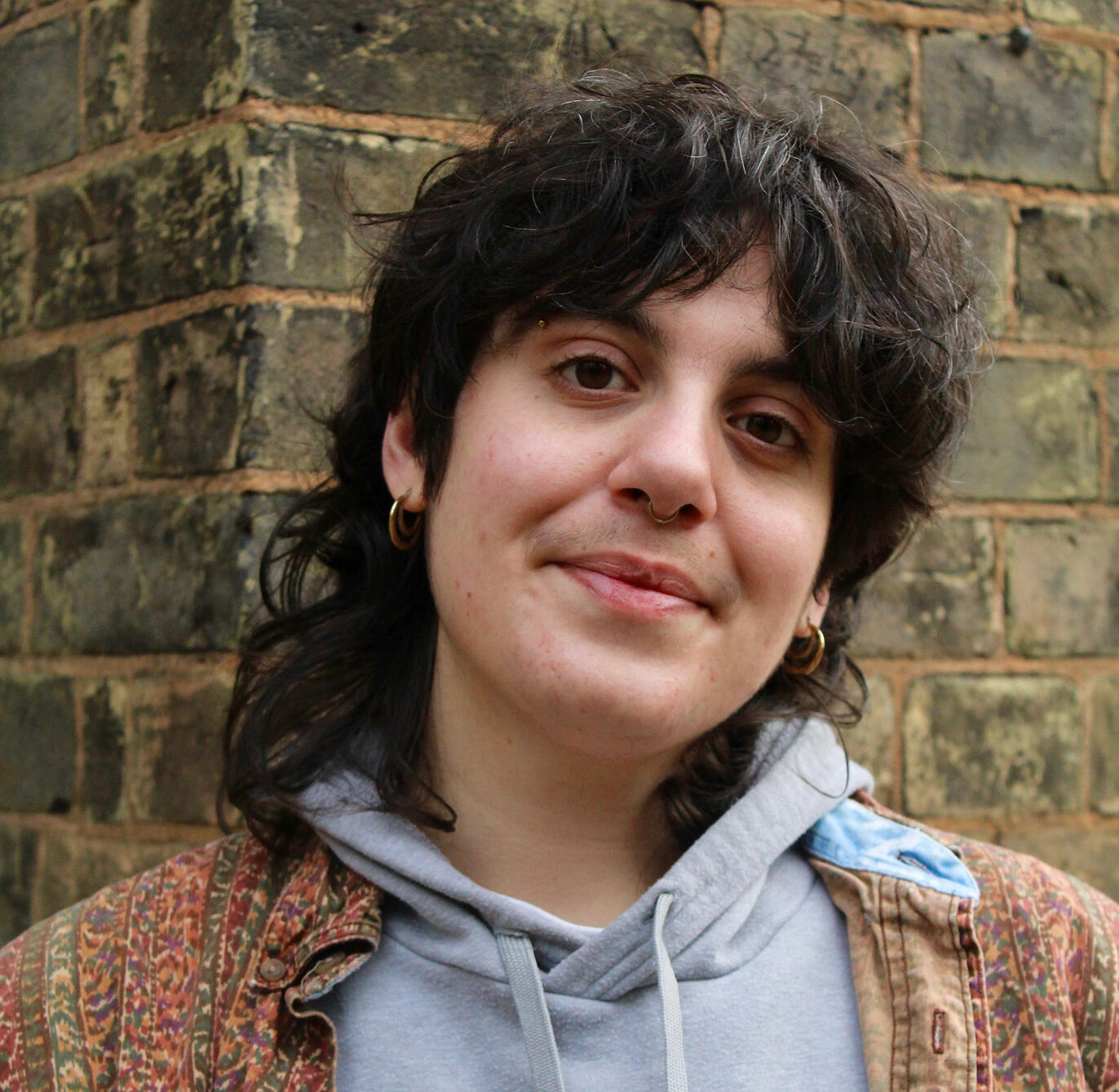 Portrait of Joey, a young, white, non binary person with dark curly hair, they are standing in front of a brick wall and gently smiling at the camera. They are wearing a grey hoodie and a multicoloured earth toned shirt over it.