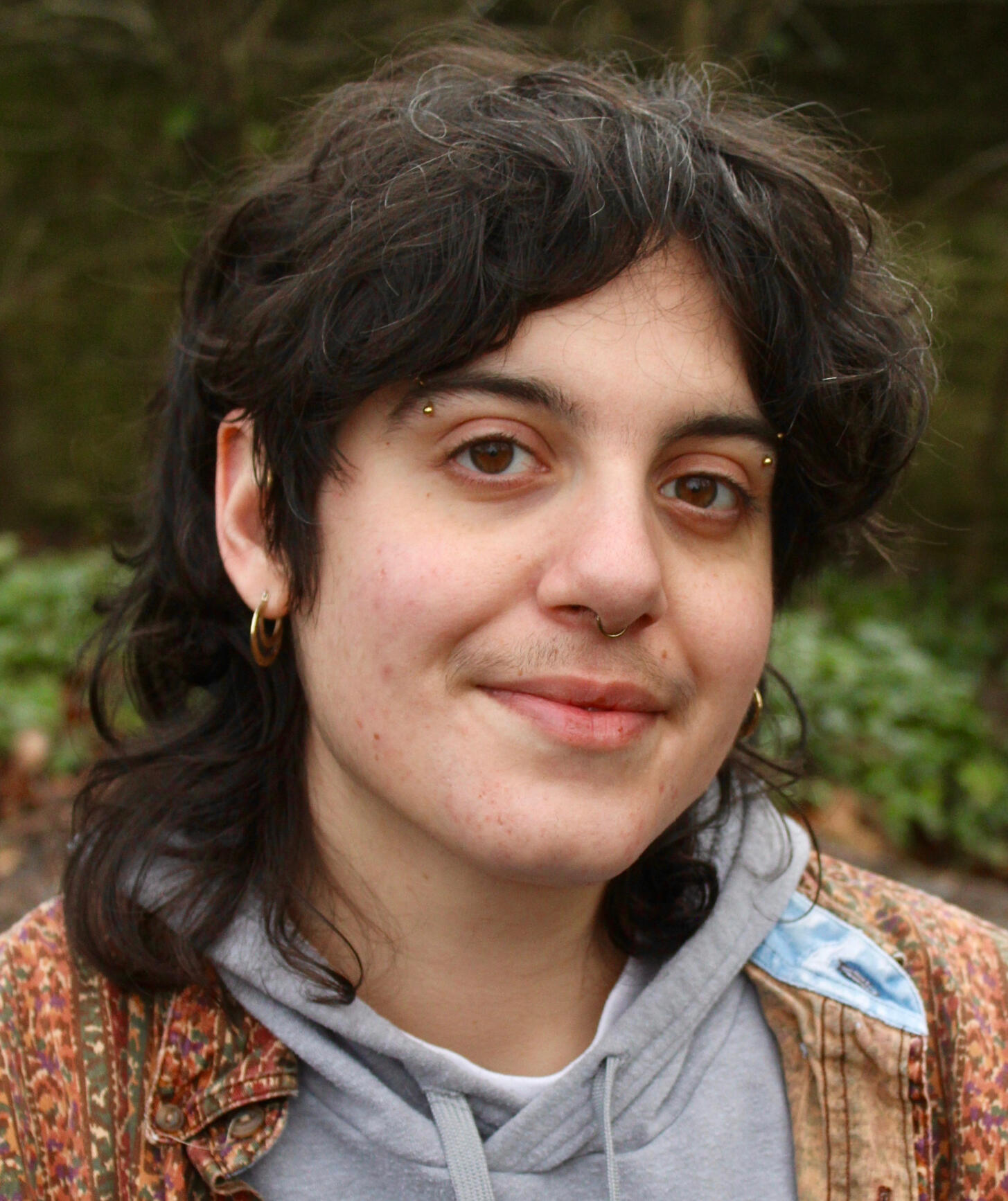 Portrait of Joey, a young, white, non binary person with dark curly hair, they are sitting in a garden in front of a brick wall and a tree.They are wearing a grey hoodie and multicoloured earth toned shirt over it. They are looking and gently smiling at th